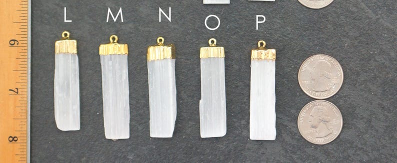 Op de afbeelding: Close-up van verschillende witte seleniet kristallen hangers met goudkleurige doppen. De hangers zijn rechthoekig en vari&euml;ren enigszins in grootte. Twee munten liggen naast de hangers ter vergelijking.