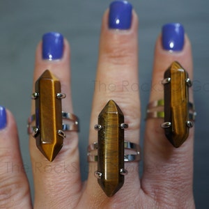 May include: Three silver rings with brown tiger's eye gemstone points. The rings are worn on the fingers of a hand with blue nail polish.