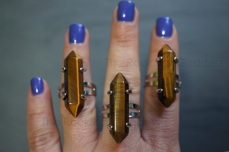 May include: Three silver rings with brown tiger's eye gemstone points. The rings are worn on the fingers of a hand with blue nail polish.