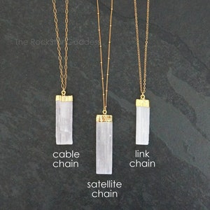 Op de afbeelding: Drie goudkleurige kettingen met rechthoekige witte hangers. De kettingen zijn gelabeld met "cable chain", "satellite chain" en "link chain". De hangers hebben goudkleurige doppen. De achtergrond is donkergrijs.
