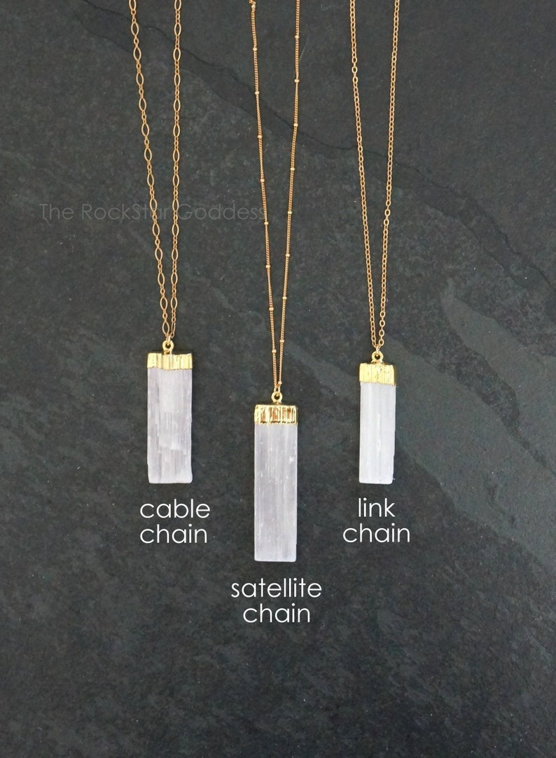 Op de afbeelding: Drie goudkleurige kettingen met rechthoekige witte hangers. De kettingen zijn gelabeld met "cable chain", "satellite chain" en "link chain". De hangers hebben goudkleurige doppen. De achtergrond is donkergrijs.