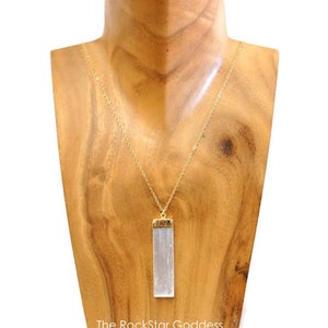 Op de afbeelding: Een goudkleurige ketting met een rechthoekige, heldere kristallen hanger. De hanger is gevat in goudkleurig metaal. De ketting wordt getoond op een houten buste. De tekst "The RockStar Goddess" staat onderaan.