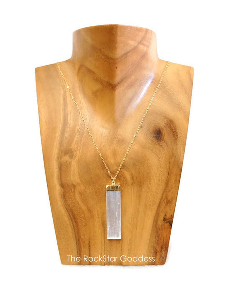 Op de afbeelding: Een goudkleurige ketting met een rechthoekige, heldere kristallen hanger. De hanger is gevat in goudkleurig metaal. De ketting wordt getoond op een houten buste. De tekst "The RockStar Goddess" staat onderaan.