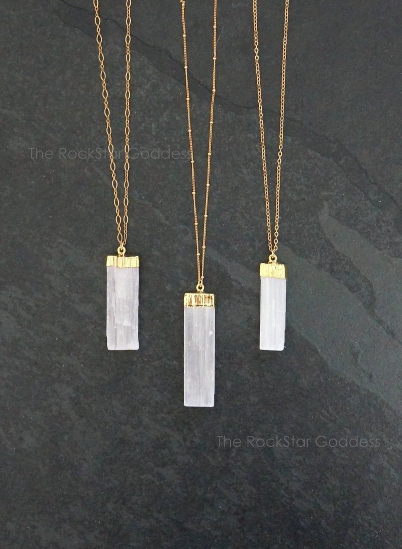 Op de afbeelding: Drie goudkleurige kettingen met rechthoekige, witte kristallen hangers. De hangers zijn afgewerkt met goudkleurig metaal. De kettingen worden gepresenteerd op een donkergrijze ondergrond. The RockStar Goddess staat op de afbeelding.