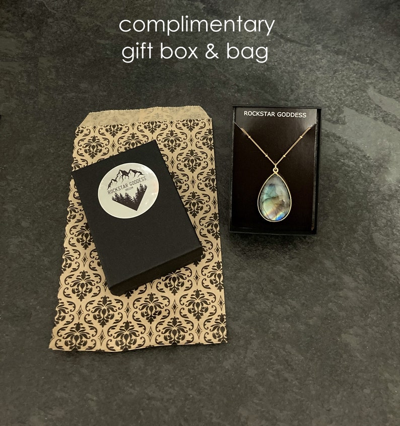 May include: A brown paper gift bag with a black and white damask pattern. The bag is next to a black gift box with a white sticker that says "Rockstar Goddess" and a mountain range graphic. The gift box is next to a small black box with a gold chain necklace with a teardrop-shaped pendant.