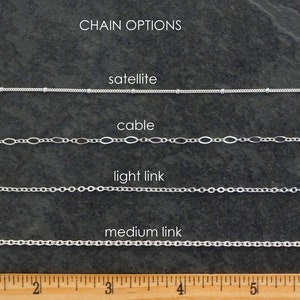 May include: Close-up of four different silver chain styles: satellite, cable, light link, and medium link. The chains are laid out horizontally on a dark gray surface with a ruler underneath for scale. The ruler is marked in inches.