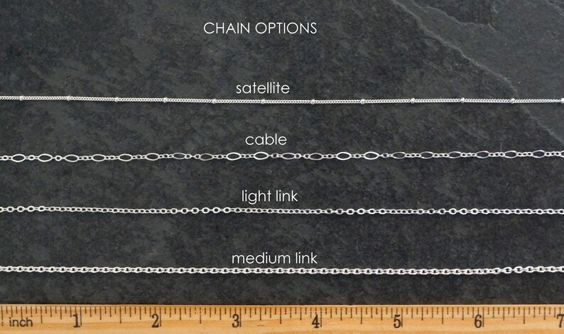 May include: Close-up of four different silver chain styles: satellite, cable, light link, and medium link. The chains are laid out horizontally on a dark gray surface with a ruler underneath for scale. The ruler is marked in inches.