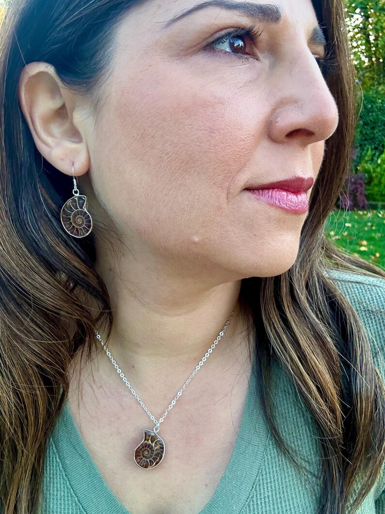 Puede incluir: Un collar de cadena de plata con un colgante de amonita marr&oacute;n. El collar lo lleva una persona con el pelo largo y casta&ntilde;o. La persona tambi&eacute;n lleva pendientes de plata con colgantes de amonita marr&oacute;n.