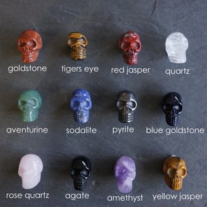 May include: Twelve different crystal skull figurines, each with a unique color and stone type. The stones include goldstone, tigers eye, red jasper, quartz, aventurine, sodalite, pyrite, blue goldstone, rose quartz, agate, amethyst, and yellow jasper.