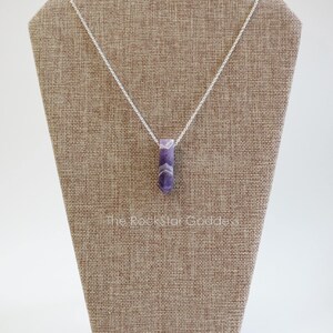 May include: A silver chain necklace with a purple amethyst pendant. The pendant is a long, thin, faceted crystal.