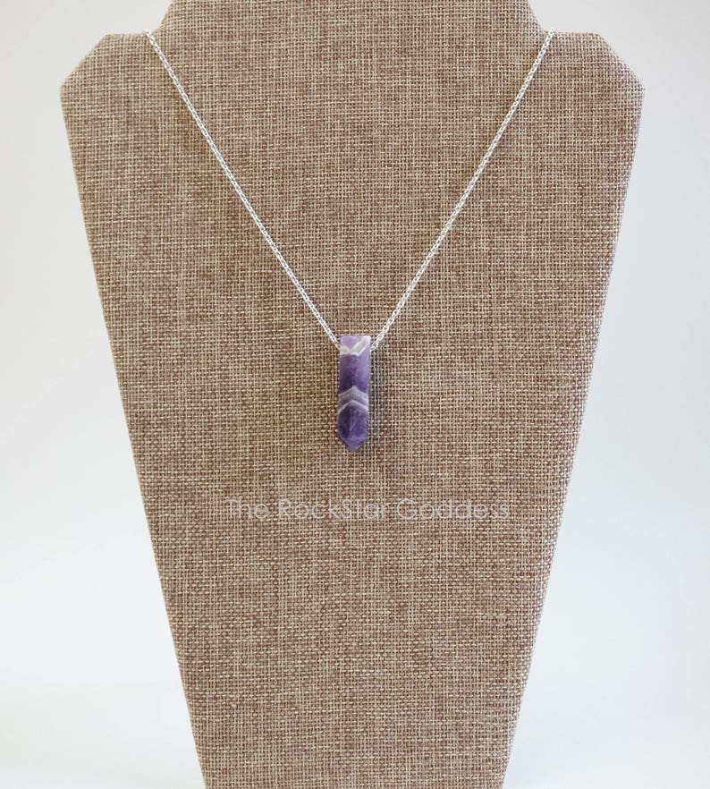 May include: A silver chain necklace with a purple amethyst pendant. The pendant is a long, thin, faceted crystal.
