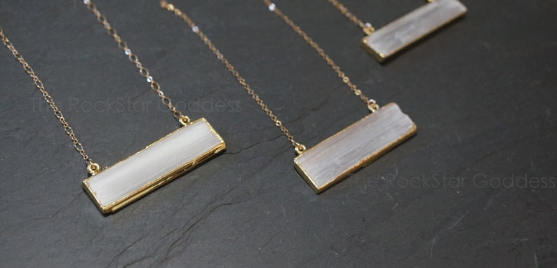 Pode incluir: Quatro colares de ouro com pingentes retangulares de cristal de selenite. Os colares s&atilde;o exibidos em uma superf&iacute;cie preta.