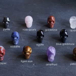 May include: A collection of 12 skull-shaped crystal carvings, each made from a different type of stone. The stones include aventurine, agate, amethyst, blue goldstone, goldstone, pyrite, quartz, red jasper, sodalite, tigers eye, and yellow jasper.