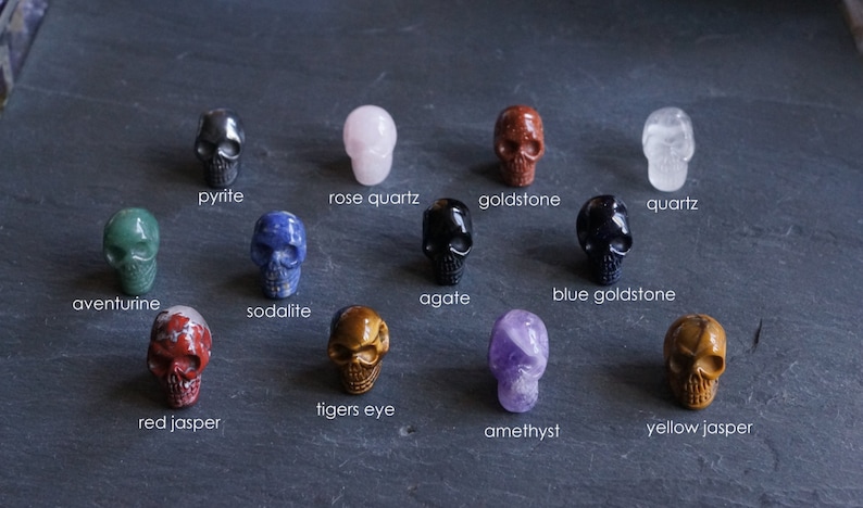 May include: A collection of 12 skull-shaped crystal carvings, each made from a different type of stone. The stones include aventurine, agate, amethyst, blue goldstone, goldstone, pyrite, quartz, red jasper, sodalite, tigers eye, and yellow jasper.