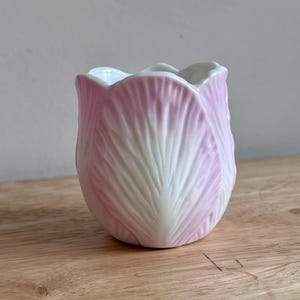 May include: A small ceramic vessel shaped like a tulip flower with a scalloped rim. The exterior features a gradient design transitioning from a soft white base to a delicate light pink color at the edges, with embossed petal textures.