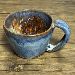 May include: A handmade ceramic mug with a blue and brown glaze. The mug has a rounded shape with a sturdy handle. The interior features a unique brown and gold pattern, and the rim is a darker brown.