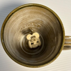 以下が含まれることがあります： 手作りのセラミックマグカップ。外側と内側は明るい茶色。小さなクリーム色のネズミの置物がマグカップの中央に配置されています。マグカップには取っ手があり、温かい飲み物にも冷たい飲み物にも適しています。