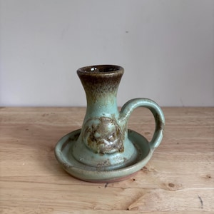 Può includere: Un portacandele in ceramica con un'ampia base circolare, un alloggiamento cilindrico centrale e un manico laterale curvo. L'oggetto presenta una smaltatura verde chiaro maculata con accenti marrone scuro sul bordo e un emblema circolare.