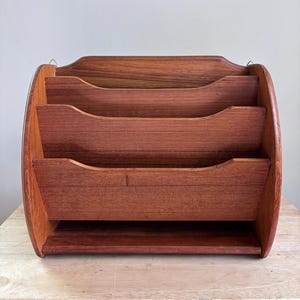 May include: A wooden desk organizer with three tiered compartments. The organizer is made of reddish-brown wood with a curved design. The top has a small handle for hanging. The compartments have a curved front for easy access.