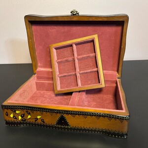 Puede incluir: Caja de joyería de madera abierta con interior de terciopelo rojo y una bandeja extraíble. La caja es marrón con diseños decorativos de hojas y puntos negros y amarillos. La caja está abierta, revelando el interior.