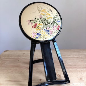 Könnte beinhalten: Ein runder Standspiegel mit schwarzem Rahmen und einer goldfarbenen, kreisförmigen Platte, die mit einer bunten Blumenillustration aus gelben, roten und blauen Blüten, grünen Blättern und hohen Gräsern verziert ist. Der Spiegel steht auf einem schwarzen Ständer.