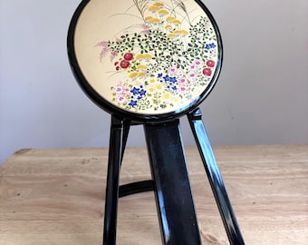 May include: A round, black-framed standing mirror featuring a gold-colored circular panel decorated with a colorful floral illustration of yellow, red, and blue flowers, green leaves, and tall grasses. The mirror is supported by a black stand.