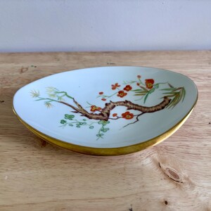 May include: An oval-shaped white porcelain dish with a gold-trimmed rim. The center features a hand-painted design of a brown tree branch with small red flowers, green leaves, and thin yellow accents, creating a delicate botanical illustration.