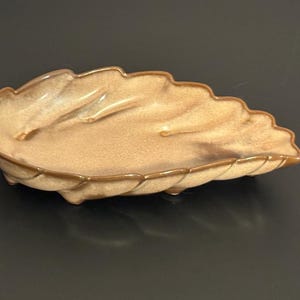 May include: A leaf-shaped ceramic dish with a light brown glaze and darker brown trim. The dish has a textured surface and a scalloped edge, resembling a large leaf. The dish is likely used for serving or display.