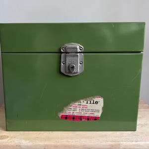 May include: A green metal file box with a silver latch and a partial sticker. The box is rectangular and sits on a wooden surface. The sticker has red and white accents and text that is partially visible.