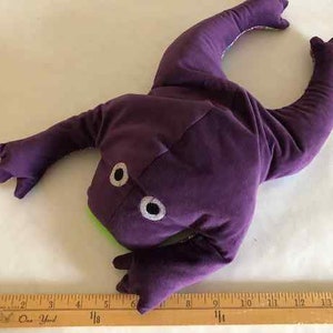 Könnte beinhalten: Ein lila Plüschfroschspielzeug mit weißen Augen und einem grünen Mund. Der Frosch liegt auf dem Rücken mit ausgestreckten Beinen.