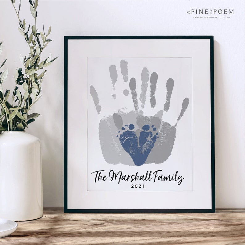 Family handprint art print with blue baby boys footprint heart. Customized with family name, colors and year. Design is displayed in a modern black frame in a farmhouse setting.