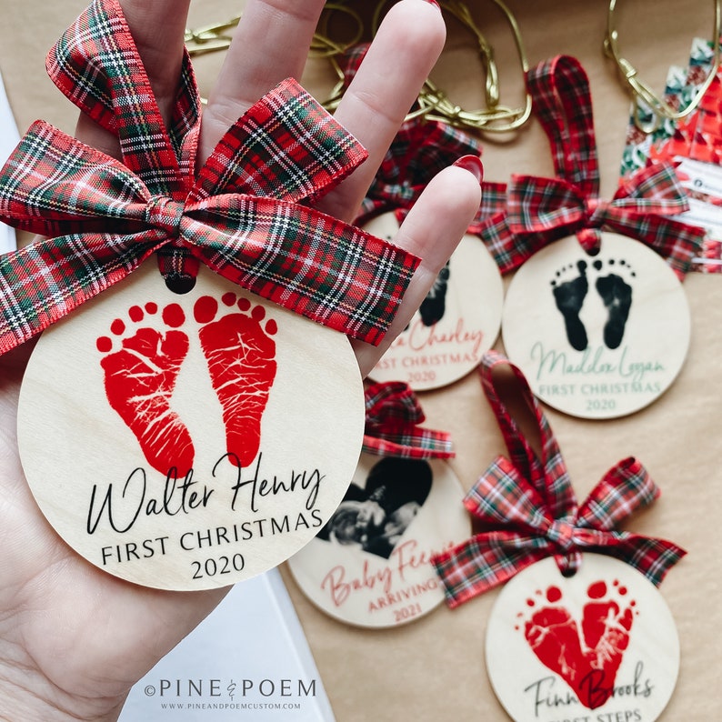 Maker holding Personalized Baby Footprint First Christmas Ornament. Wood ornament with plaid ribbon, baby name and date.