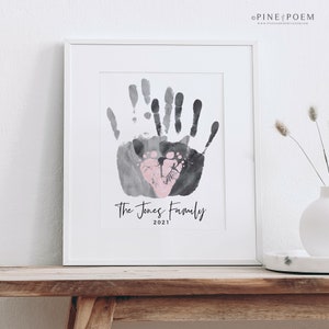 Family handprint art print with pink baby footprint heart. Customized with family name, colors and year. Design is displayed in a white frame in a modern farmhouse setting.