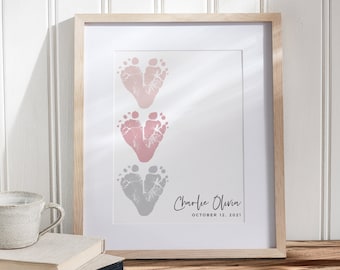 May include: A framed print with three sets of baby footprints in the shape of hearts. The top two sets are pink and the bottom set is gray. The print includes the name "Charlie Olivia" and the date "October 12, 2021".