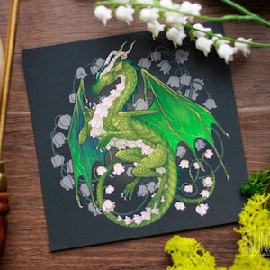 May include: A square print featuring a green dragon with white accents and large green wings, surrounded by white flowers, set against a dark background. The print is on a wooden surface, with the text "Sunset Dragon" visible.