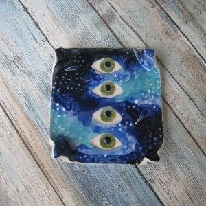 Puede incluir: Un azulejo de cerámica cuadrado con fondo azul y blanco y cuatro ojos verdes pintados sobre él.