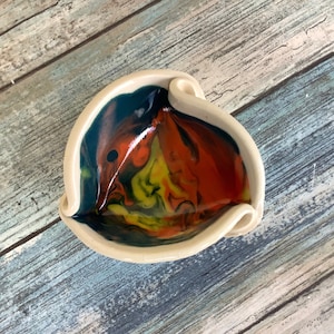 May include: A small, white ceramic bowl with a unique, tri-lobed shape. The bowl's interior is painted with a swirling pattern of blue, yellow, orange, and red.
