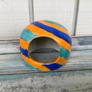 May include: A ceramic dome-shaped hide with a large opening. The hide is painted with orange, blue, and green stripes.