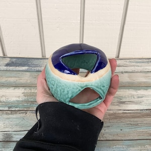 May include: A ceramic decorative object with a rounded shape and a hole in the center. The object features a color scheme of dark blue, light blue, beige, and teal. The surface has a textured, dimpled pattern.