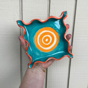 Peut inclure: Un plat en céramique turquoise avec un bord ondulé et un motif de cercle concentrique orange et blanc.