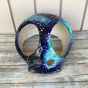 May include: A ceramic skull planter with a galaxy design in shades of blue, green, and white. The skull has two large eye sockets and a smaller mouth opening.