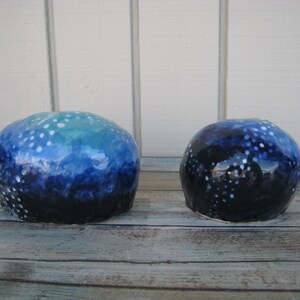 May include: Two ceramic spheres with a blue and white speckled glaze. The larger sphere has a light blue and white glaze, while the smaller sphere has a darker blue and white glaze.