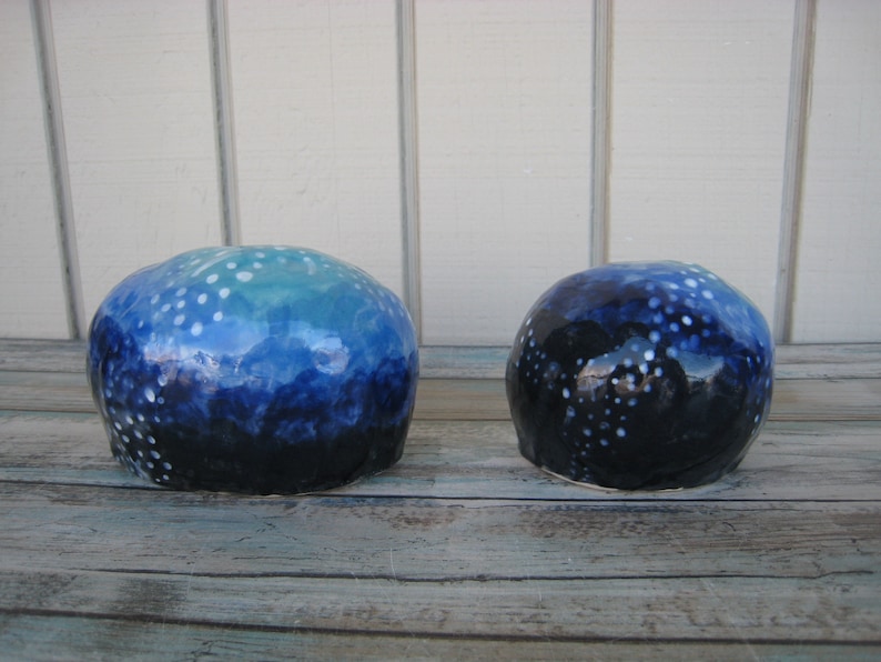 May include: Two ceramic spheres with a blue and white speckled glaze. The larger sphere has a light blue and white glaze, while the smaller sphere has a darker blue and white glaze.