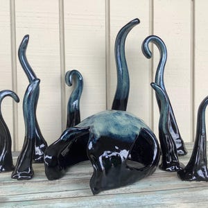 May include: A ceramic sculpture with a dark blue and black glaze. The sculpture features a central, rounded form with several curved, horn-like appendages of varying heights. The piece is displayed on a weathered, light blue wooden surface.