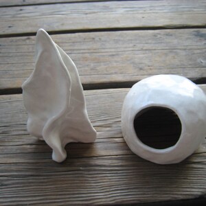 May include: Two white ceramic sculptures. One is a tall, abstract shape with a smooth, flowing form. The other is a round, hollow shape with a hole in the center.