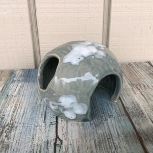 May include: A gray ceramic hide with white glaze, featuring a rounded shape and two openings. This hide is designed for small animals like reptiles or amphibians.