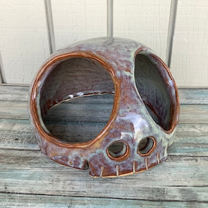 May include: A ceramic skull planter with a brown and green glaze. The skull has two eye holes and a mouth hole. The planter is sitting on a wooden surface.