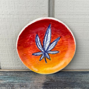 May include: A small ceramic dish with a sunset gradient design and a blue and white marijuana leaf illustration.