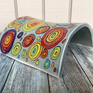 Puede incluir: Un arco de cerámica con un esmalte azul claro y un patrón colorido de círculos concéntricos en tonos de rojo, amarillo, verde y azul.