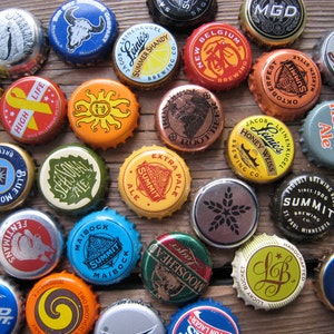 May include: A collection of colourful bottle caps with various brewery logos and names, including "Leinie's", "Summit", "Schlafly", "Two Hearted Ale", "Bud Light", and "Moosehead".
